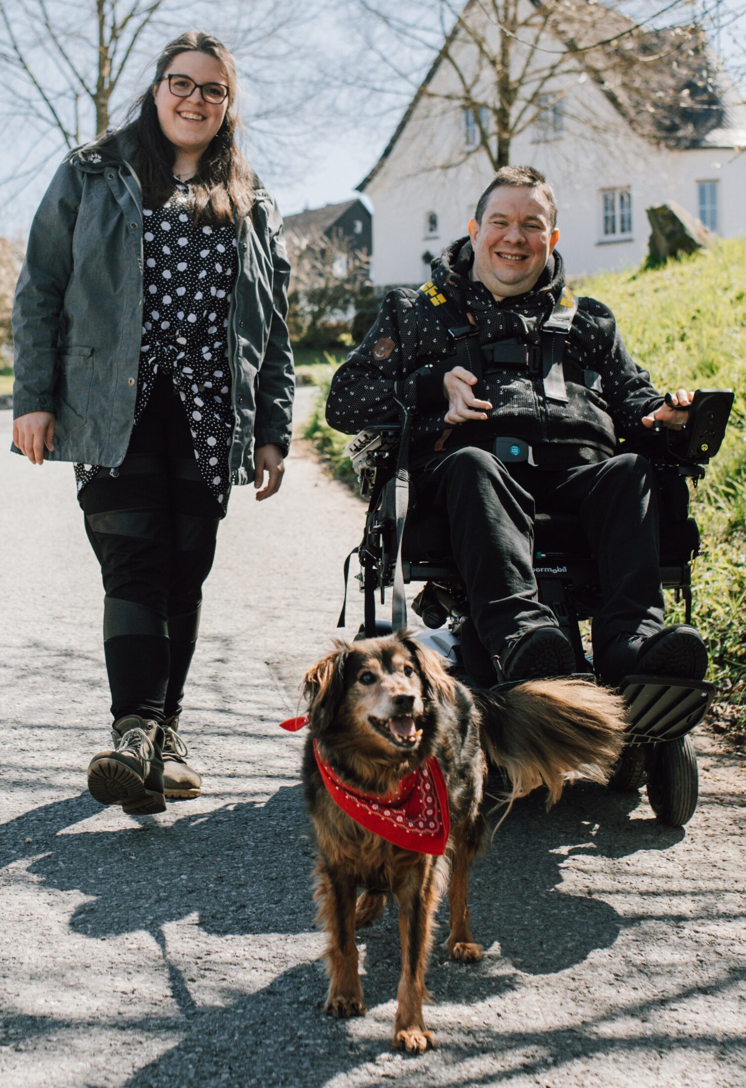 Eine lächelnde Frau geht neben einem Mann im Elektrorollstuhl spazieren, der einen Hund mit rotem Halstuch an der Leine führt, im Hintergrund ein Haus und sonniges Frühlingswetter.