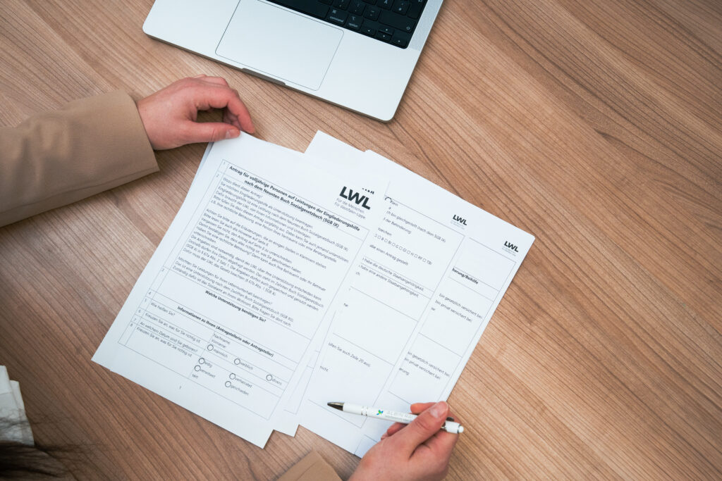 Zwei Personen sitzen an einem Holztisch und füllen gemeinsam ein Antragsformular der LWL (Landschaftsverband Westfalen-Lippe) für Eingliederungshilfe aus, ein Laptop steht daneben.