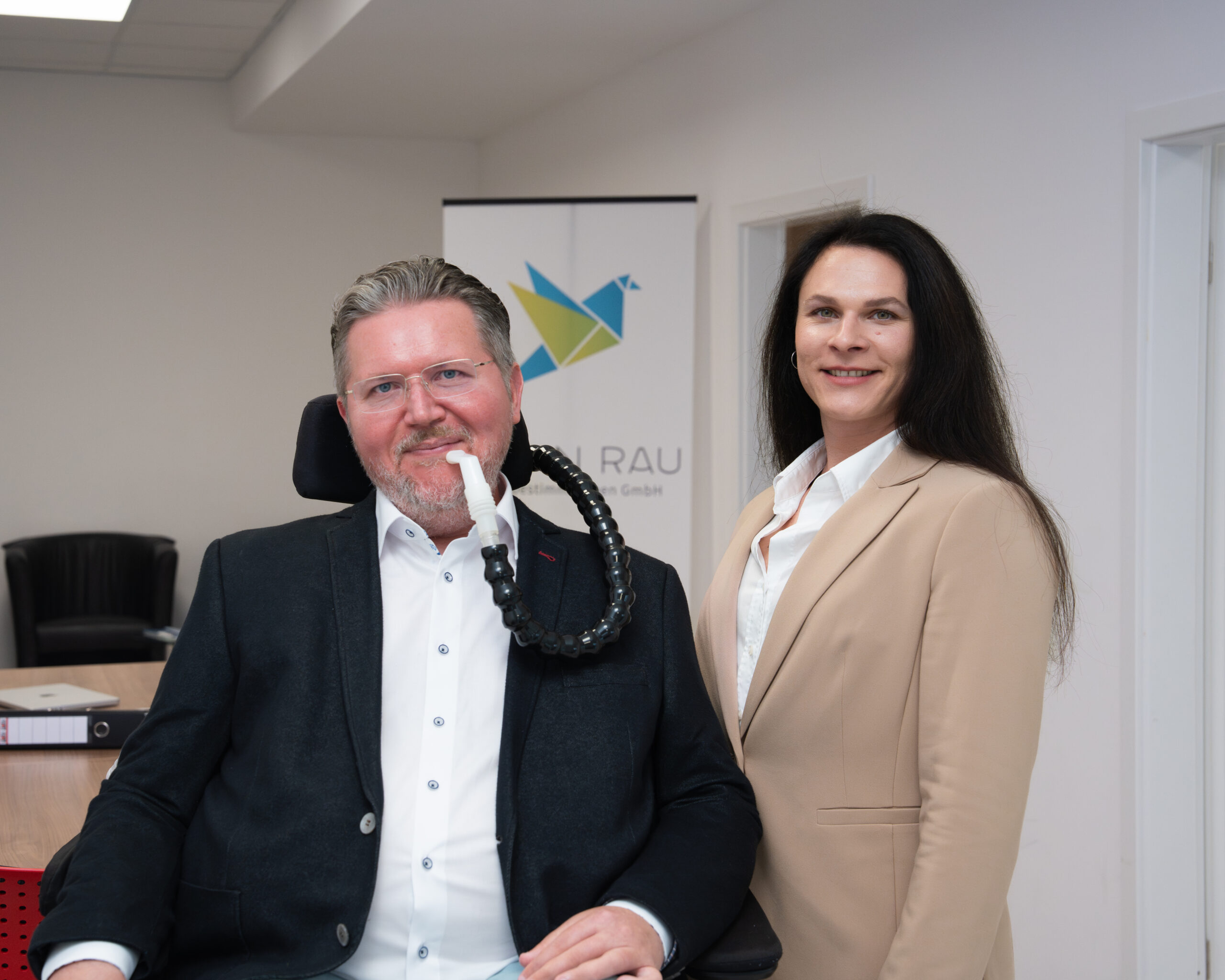 Ein Mann im Rollstuhl mit Kinnsteuerung und eine Frau in beigem Blazer stehen lächelnd nebeneinander in einem Büroraum, im Hintergrund ein Aufsteller mit dem Logo "Albin Rau".