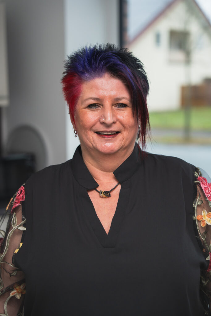 Eine lächelnde Frau mit rot-blau gefärbten Haaren und Headset sitzt im Büro vor einem Computer, im Hintergrund Pflanzen und Arbeitsplätze.