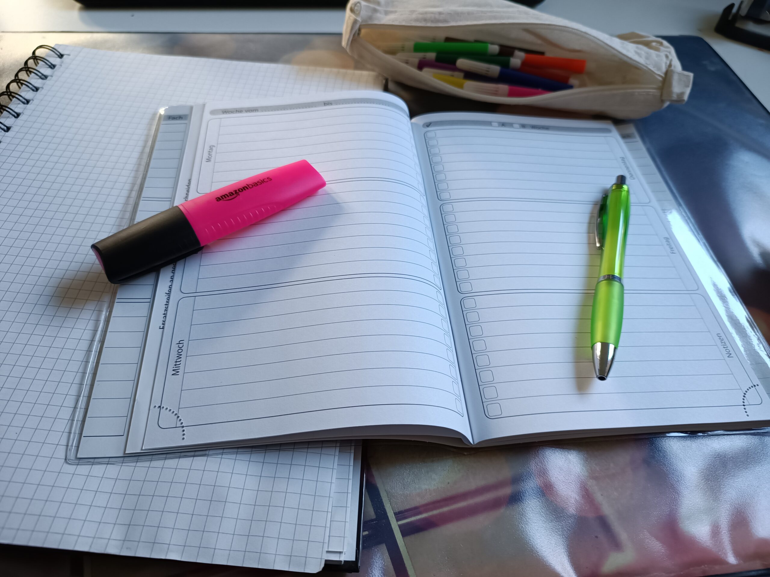 Aufgeschlagener Wochenplaner mit Checklisten-Spalten, pinkem Textmarker und grünem Kugelschreiber, daneben ein Stiftemäppchen mit bunten Filzstiften.